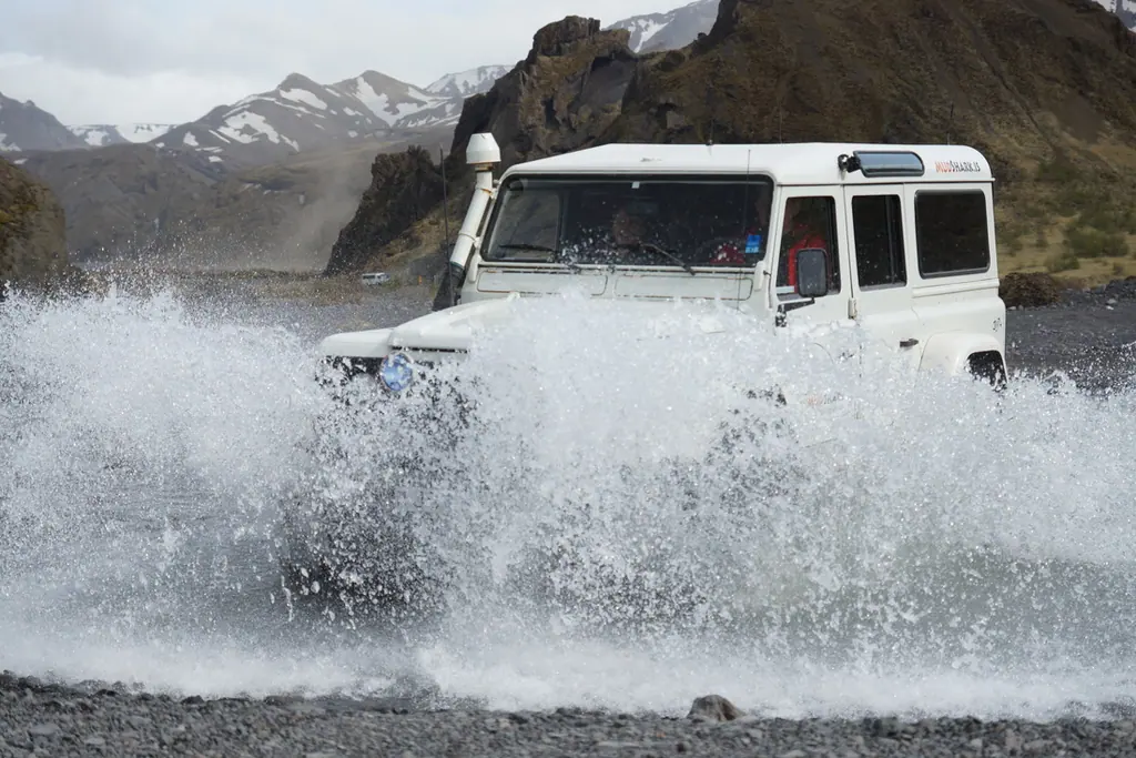 Hvítur jeppi ekur yfir jökulá og þeytir upp vatnsvegg.