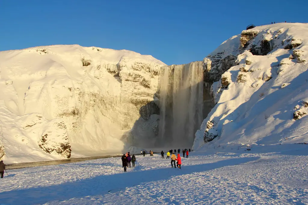 Breiður foss fellur á milli snævi þakinna hlíða, og röð af fólki í litríkum úlpum stendur smá við rætur hans.